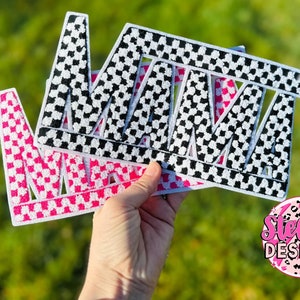 May include: Embroidered patches spelling "MAMA" in a bold, block letter style. One patch features a pink and white checkered pattern, while the other has a black and white checkered design. The patches are held against a blurred green background.