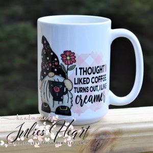 May include: White ceramic coffee mug with a pink and white checkered background. A cartoon gnome with a black hat and a pink flower is holding a cup of coffee. The text on the mug reads "I thought I liked coffee. Turns out, I like creamer."