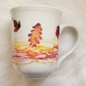 May include: White ceramic mug with a fall-themed design. The mug features a watercolor-like background with shades of pink, orange, and yellow. Three autumn leaves are depicted in shades of red and brown.