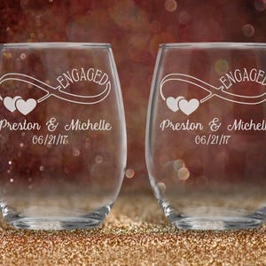 May include: Two clear glass wine glasses with the text "Engaged" and an infinity symbol with two hearts. The glasses are personalized with the names "Preston & Michelle" and the date "06/21/17".