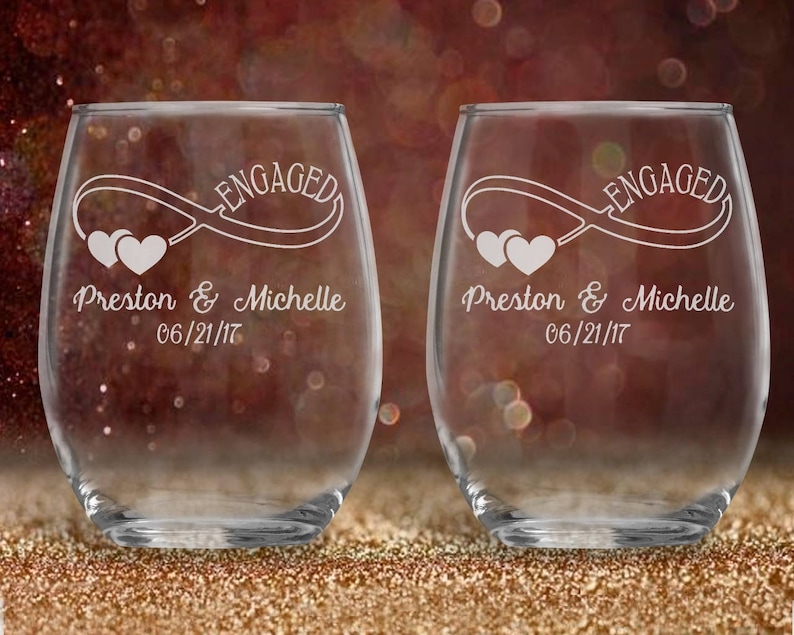 May include: Two clear glass wine glasses with the text "Engaged" and an infinity symbol with two hearts. The glasses are personalized with the names "Preston & Michelle" and the date "06/21/17".