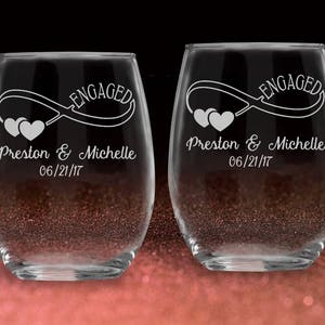 May include: Two clear glass wine glasses with the word "Engaged" and an infinity symbol with hearts engraved on them. The glasses are personalized with the names "Preston & Michelle" and the date "06/21/17".