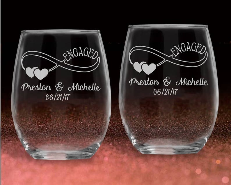 May include: Two clear glass wine glasses with the word "Engaged" and an infinity symbol with hearts engraved on them. The glasses are personalized with the names "Preston & Michelle" and the date "06/21/17".