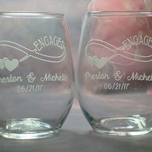 May include: Two clear glass wine glasses with the word "ENGAGED" etched on the front. The glasses also have an infinity symbol with two hearts and the text "Preston & Michelle 06/21/17" etched on the front.