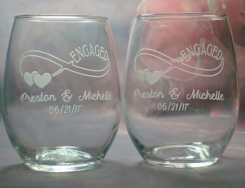 May include: Two clear glass wine glasses with the word "ENGAGED" etched on the front. The glasses also have an infinity symbol with two hearts and the text "Preston & Michelle 06/21/17" etched on the front.
