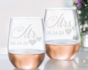 May include: Two clear glass wine glasses with the words "Mr." and "Mrs." etched on them. The glasses also have the date "05-24-20" and three small hearts etched on them. The glasses are filled with a light pink liquid.