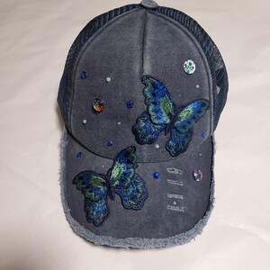 May include: A denim baseball cap with a mesh back, adorned with embroidered blue and green butterflies and scattered rhinestones. The cap has a distressed brim and a button on top.