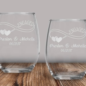 May include: Two clear glass wine glasses with an infinity symbol and the words "Engaged" and "Preston & Michelle 06/21/17" etched on the front.