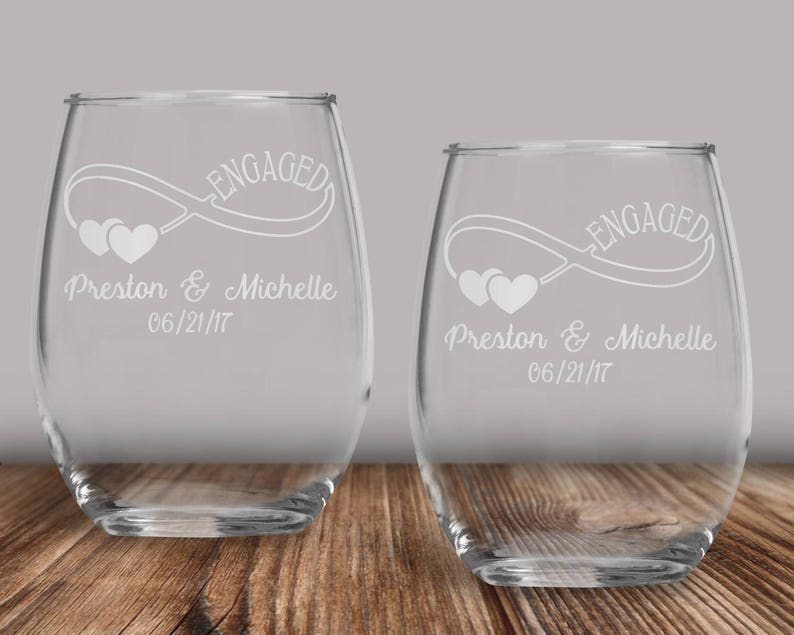 May include: Two clear glass wine glasses with an infinity symbol and the words "Engaged" and "Preston & Michelle 06/21/17" etched on the front.