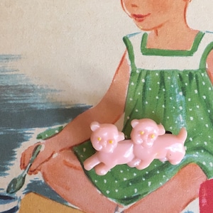 May include: Illustration of two young girls playing with toy furniture and pink bear figurines. One girl wears a green polka-dot dress, and the other wears a pink dress. A small table and chairs are also present.