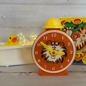 May include: Orange vintage tiger-themed alarm clock with a yellow hat-shaped top. The clock face features a tiger illustration and yellow hands. A yellow rubber duck sits in a small white bathtub with fairy lights.