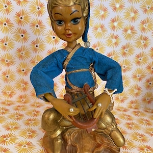 Op de afbeelding: Een beeldje van een zittend figuur met blond haar, een blauwe muts en een blauw shirt met lange mouwen en gouden randen. Het figuur houdt een kleine bruine lier vast en draagt een goudkleurige broek met parelachtige accenten op de handen.