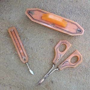 Puede incluir: Un juego de manicura vintage de color melocotón con detalles plateados. El juego incluye una lima de uñas, unas tijeras y un empujador de cutículas. Las piezas tienen un diseño decorativo y están dispuestas sobre una superficie neutra.