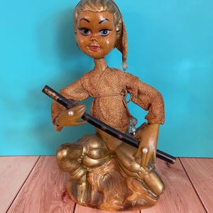 May include: A decorative figurine of a person with a wood-grain finish, wearing a textured copper-coloured top and matching trousers. The figure has a pointed cap, holds a long black rod, and features small white pearl-like accents on the hands and clothing.