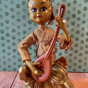 May include: A vintage-style figurine of a seated person with a wood-toned finish, wearing a textured metallic gold top and a matching cap. The figure holds a brown stringed musical instrument and is adorned with small white pearl-like accents.