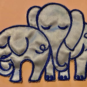 Könnte beinhalten: Ein Stoffaufnäher mit zwei gestickten Elefanten in hellem Silbergrau, die mit dunkelblauem Garn umrandet sind. Der größere Elefant steht auf der rechten Seite, während sich ein kleinerer Elefant links daneben befindet.