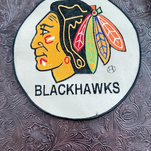 May include: A circular embroidered patch featuring the Chicago Blackhawks logo on a cream-colored background. The design depicts a profile of a Native American figure with black hair and colorful feathers in red, green, blue, and orange, with the text BLACKHAWKS.