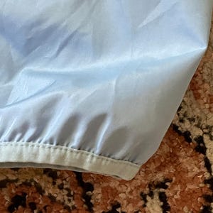 K&ouml;nnte beinhalten: Eine hellblaue Jacke mit einem schwarzen Rei&szlig;verschluss und einem schwarzen Saum am unteren Rand.