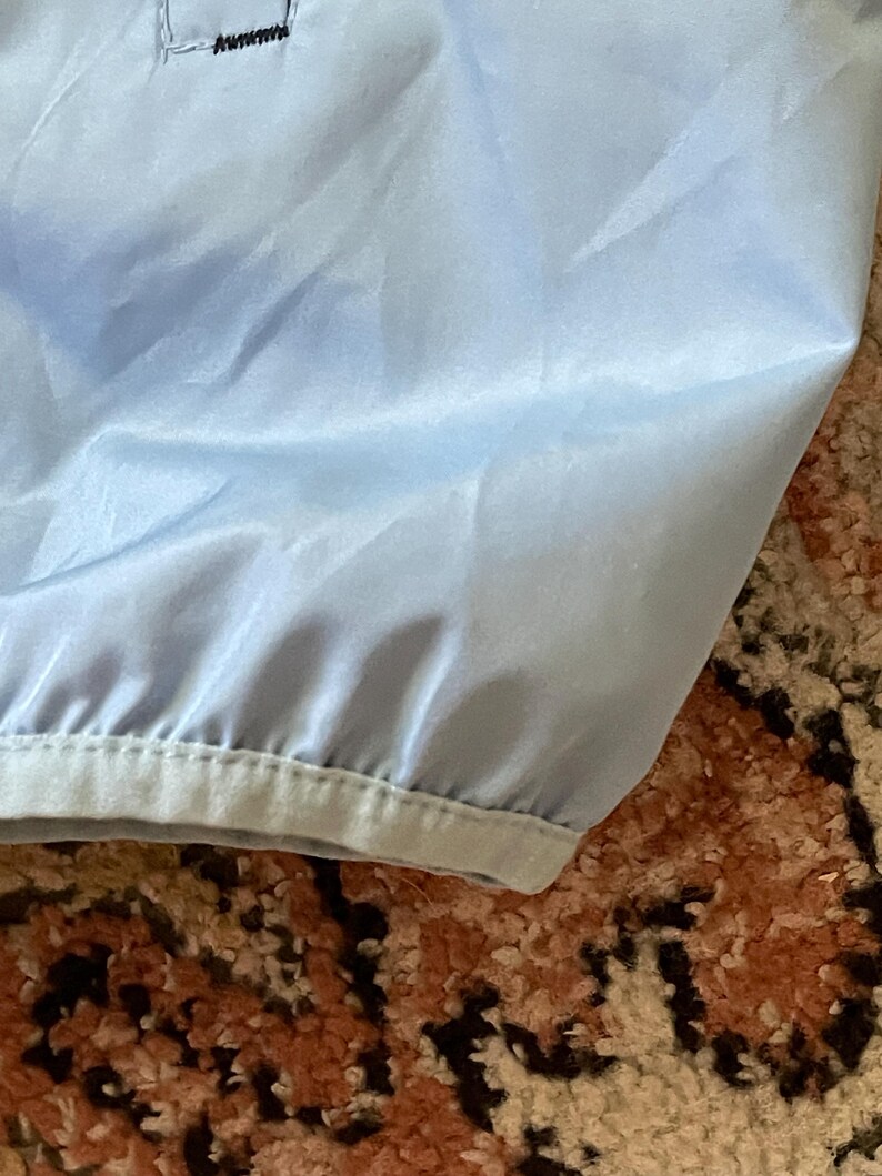 K&ouml;nnte beinhalten: Eine hellblaue Jacke mit einem schwarzen Rei&szlig;verschluss und einem schwarzen Saum am unteren Rand.