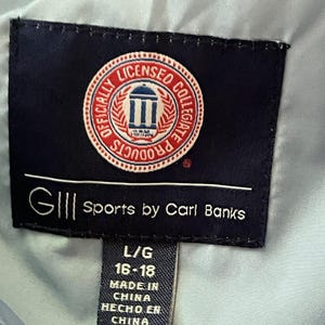 K&ouml;nnte beinhalten: Ein schwarzes und wei&szlig;es Etikett mit dem Text "GIII Sports by Carl Banks" und ein kleineres Etikett mit dem Text "L/G 16-18 MADE IN CHINA HECHO EN CHINA". Das gr&ouml;&szlig;ere Etikett hat ein rotes, wei&szlig;es und blaues kreisf&ouml;rmiges Logo mit einem Geb&auml;ude und dem Text "OFFICIALLY LICENSED COLLEGIATE PRODUCTS".
