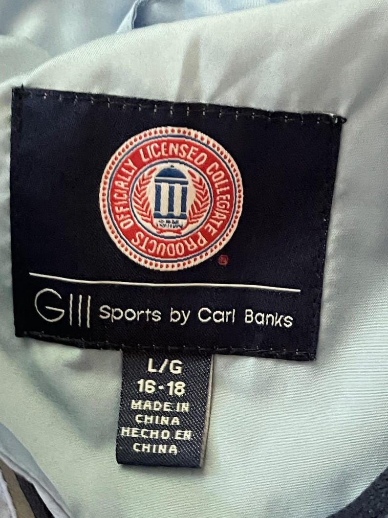K&ouml;nnte beinhalten: Ein schwarzes und wei&szlig;es Etikett mit dem Text "GIII Sports by Carl Banks" und ein kleineres Etikett mit dem Text "L/G 16-18 MADE IN CHINA HECHO EN CHINA". Das gr&ouml;&szlig;ere Etikett hat ein rotes, wei&szlig;es und blaues kreisf&ouml;rmiges Logo mit einem Geb&auml;ude und dem Text "OFFICIALLY LICENSED COLLEGIATE PRODUCTS".