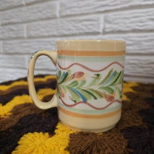 May include: A ceramic mug with a cream-colored base featuring horizontal orange and light green stripes. The center band displays a hand-painted pattern of green leaves and blue accents framed by wavy pink lines. The handle is curved and cream-colored.