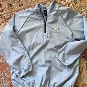 K&ouml;nnte beinhalten: Eine hellblaue Windbreaker-Jacke mit Rei&szlig;verschluss und einem grau-wei&szlig;en Muster. Die Jacke hat ein "Tar Heels"-Logo mit einem Carolina-blauen "N" auf der Vorderseite.