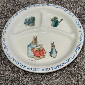 May include: A white ceramic divided plate featuring Peter Rabbit illustrations. The rim has a blue vine pattern. The center shows Peter Rabbit and friends, with a watering can and Peter in a garden. Text reads PETER RABBIT AND FRIENDS.