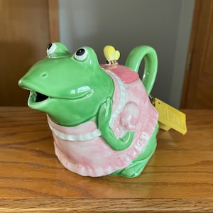 May include: A ceramic teapot shaped like a green frog wearing a pink dress. The frog has large eyes and an open mouth. A yellow butterfly sits on the lid. The teapot is on a wooden surface.