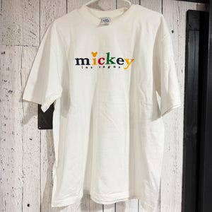 May include: A white short-sleeve t-shirt featuring an embroidered logo on the chest. The text reads mickey in multi-colored letters with a yellow Mickey Mouse head icon replacing the letter i, and the words las vegas printed in black below.