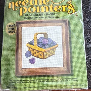 May include: A vintage needlepoint kit titled "Blackberry Basket" by Nancy Overton. The kit features a framed cross-stitch design of a yellow basket filled with purple berries. The packaging is green with the words "Needle Pointers" in yellow.