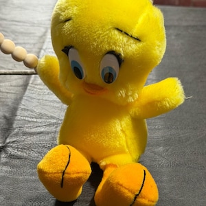 May include: A plush Tweety Bird toy featuring bright yellow fur, large blue eyes with black eyelashes, and orange feet. The character is attached to a string of small, round, light-colored wooden beads that curve around the top of the head.