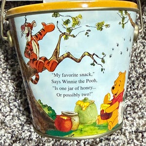 May include: A metal bucket with a yellow rim and a silver handle. The bucket features illustrations of Winnie the Pooh and Tigger, with text that reads "My favorite snack, Says Winnie the Pooh, Is one jar of honey... Or possibly two!"