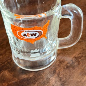 May include: Clear glass shot glass with a handle. The glass has a textured surface and features an orange and brown A&W logo in the shape of the United States. The shot glass is empty and sits on a wooden surface.