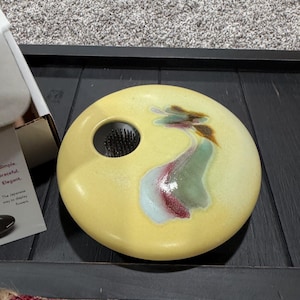 May include: A round, yellow ceramic flower frog base with a smooth, glossy finish and a central circular opening containing a metal pin holder. The surface features an abstract, blended glaze pattern in shades of green, brown, and maroon.