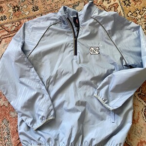 K&ouml;nnte beinhalten: Hellblaue Windbreaker-Jacke mit Rei&szlig;verschluss und grau-wei&szlig;em Muster. Die Jacke hat ein kleines gesticktes Logo mit der Aufschrift "Tar Heels" und zeigt ein Carolina-blaues "N" mit wei&szlig;em Umriss.