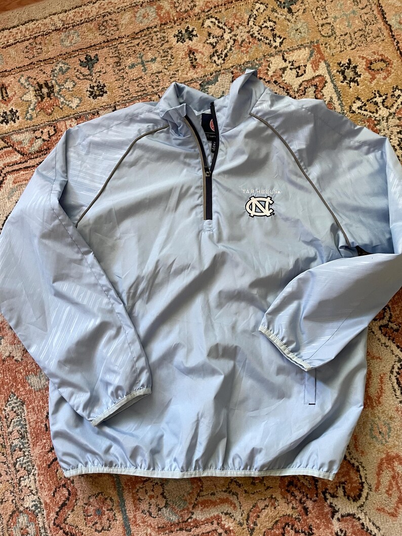 K&ouml;nnte beinhalten: Hellblaue Windbreaker-Jacke mit Rei&szlig;verschluss und grau-wei&szlig;em Muster. Die Jacke hat ein kleines gesticktes Logo mit der Aufschrift "Tar Heels" und zeigt ein Carolina-blaues "N" mit wei&szlig;em Umriss.