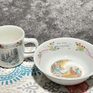 May include: A white ceramic mug and bowl set. The mug and bowl feature illustrations of a rabbit in a blue coat and other characters. The bowl has the text "For your Christening" and floral accents. The set is on a patterned surface.