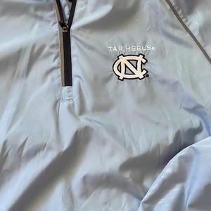 K&ouml;nnte beinhalten: Eine hellblaue Windbreaker-Jacke mit Rei&szlig;verschluss und einem wei&szlig;en gestickten Tar Heels-Logo mit einem Carolina-blauen "N" auf der Vorderseite.