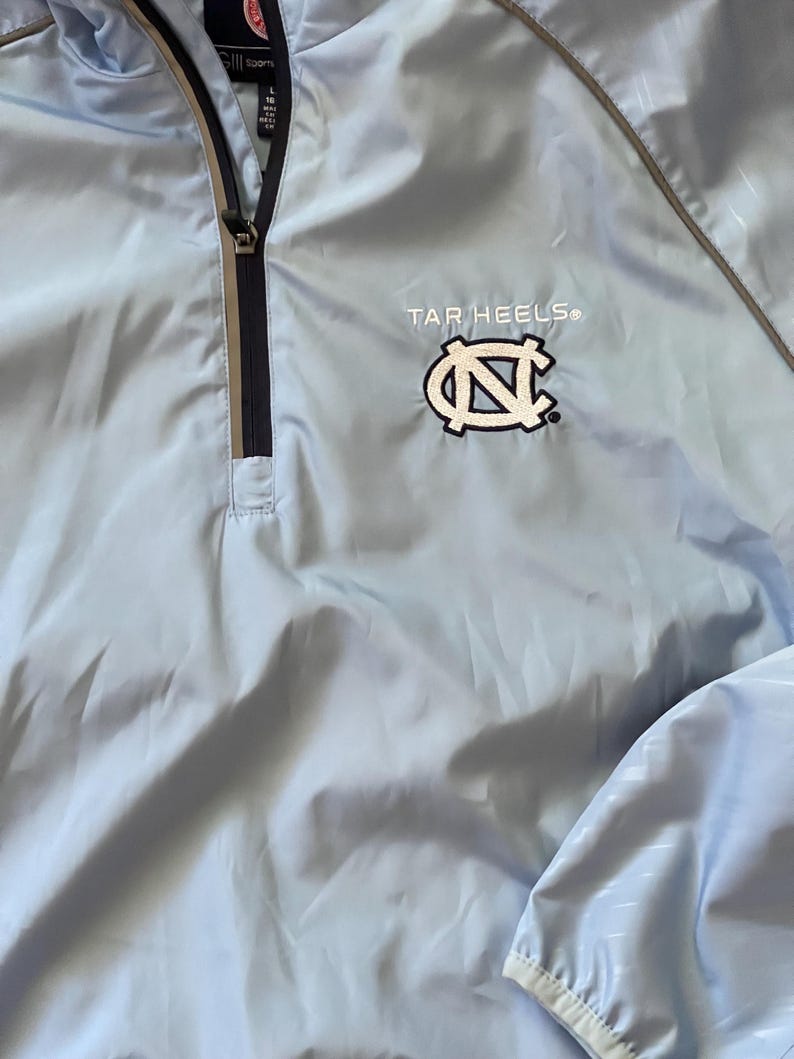 K&ouml;nnte beinhalten: Eine hellblaue Windbreaker-Jacke mit Rei&szlig;verschluss und einem wei&szlig;en gestickten Tar Heels-Logo mit einem Carolina-blauen "N" auf der Vorderseite.