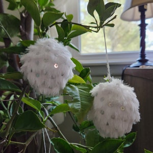 May include: Two white fluffy pom-pom ornaments with silver sequins hanging from a green leafy plant.