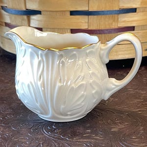 May include: White porcelain creamer with a gold rim and a leaf design. The creamer is sitting on a brown surface.