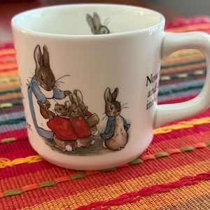 May include: A white ceramic mug featuring illustrations of Peter Rabbit and other rabbits in blue jackets and red capes. The exterior displays text snippets, and a small rabbit head is visible on the inner rim. The mug has a curved handle and a smooth finish.