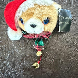 May include: A plush teddy bear head wearing a red and white Santa hat and a plaid bow tie. The bear has blue eyes and long eyelashes. Two small gold bells hang from a red string. A tag reads "Especially for you."