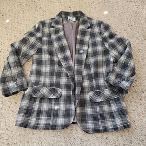 May include: A plaid blazer in shades of black, gray, and white. The jacket features a classic notched collar, two front pockets, and buttoned cuffs. The blazer is open, revealing a light purple lining.