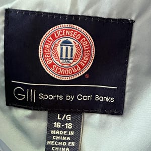K&ouml;nnte beinhalten: Ein schwarzes und blaues Etikett mit wei&szlig;em Text, der "GIII Sports by Carl Banks" und "Officially Licensed Collegiate Products" lautet. Das Etikett enth&auml;lt auch ein rotes, wei&szlig;es und blaues kreisf&ouml;rmiges Logo mit einem Geb&auml;ude und dem Text "Licensed Collegiate Products". Unter dem Etikett befindet sich ein schwarzes Etikett mit wei&szlig;em Text, der "L/G 16-18 Made in China Hecho en China" lautet.
