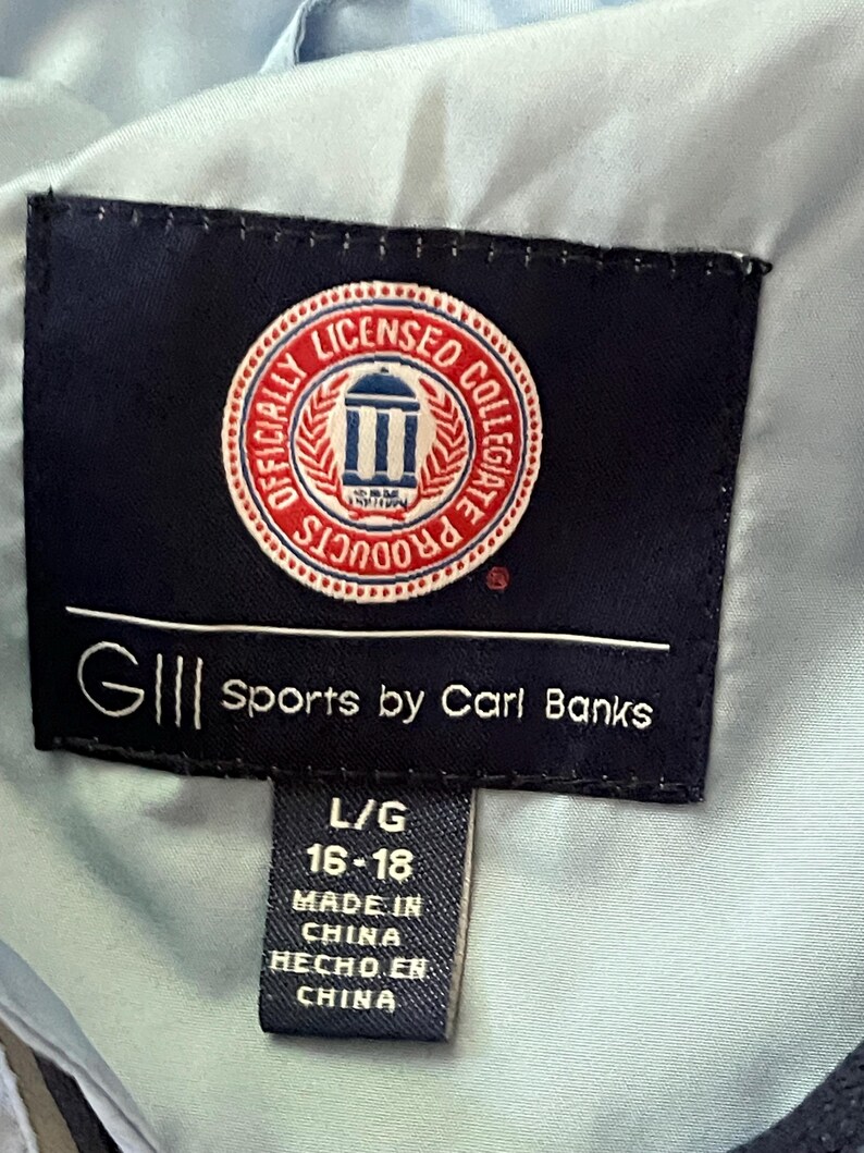 K&ouml;nnte beinhalten: Ein schwarzes und blaues Etikett mit wei&szlig;em Text, der "GIII Sports by Carl Banks" und "Officially Licensed Collegiate Products" lautet. Das Etikett enth&auml;lt auch ein rotes, wei&szlig;es und blaues kreisf&ouml;rmiges Logo mit einem Geb&auml;ude und dem Text "Licensed Collegiate Products". Unter dem Etikett befindet sich ein schwarzes Etikett mit wei&szlig;em Text, der "L/G 16-18 Made in China Hecho en China" lautet.