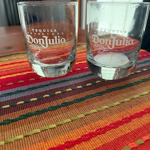 May include: Two clear glass shot glasses featuring the white printed text Tequila Don Julio. Each glass has a thick, weighted base and a cylindrical shape. The glasses are transparent with smooth rims and are positioned side by side.