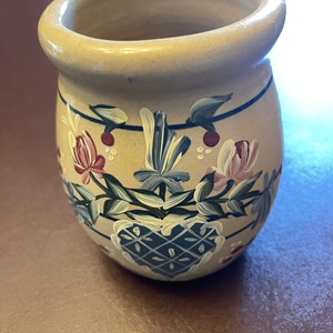 May include: A hand-painted ceramic jar with a heart design and floral accents in shades of blue, pink, and green. The jar has a beige background and a slightly rounded shape.