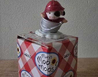 May include: A figurine of a cow wearing a red hat and shirt, sitting in a silver bucket, atop a box. The box is red and white checkered with the words "Mary's Moo Moos" and a sunflower design.